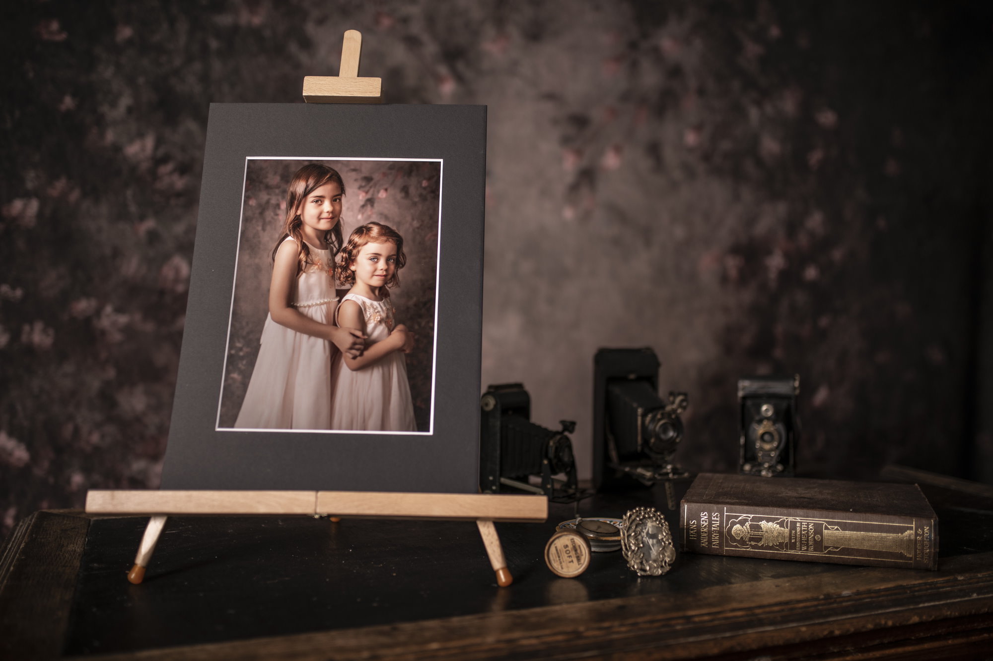 Easel with an image on, sat on a desk with vintage objects surrounding it. Including old camera, a small photo frame and a book, sat on a desk, in front of a floral backdrop.