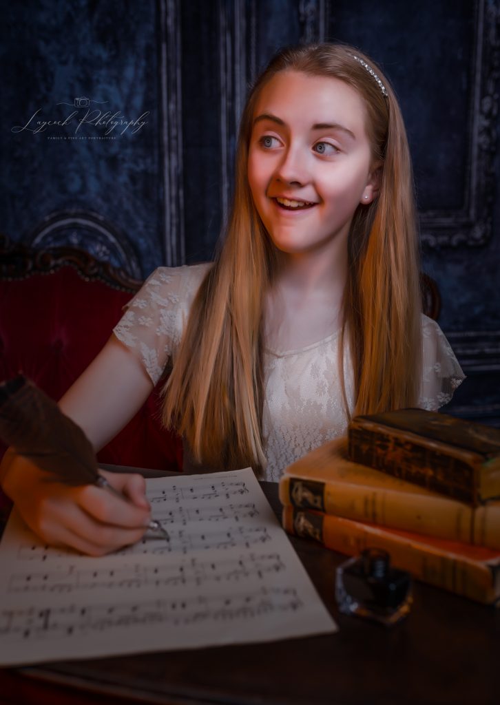 young girl at writing desk holding a quill. Books and music score on table.