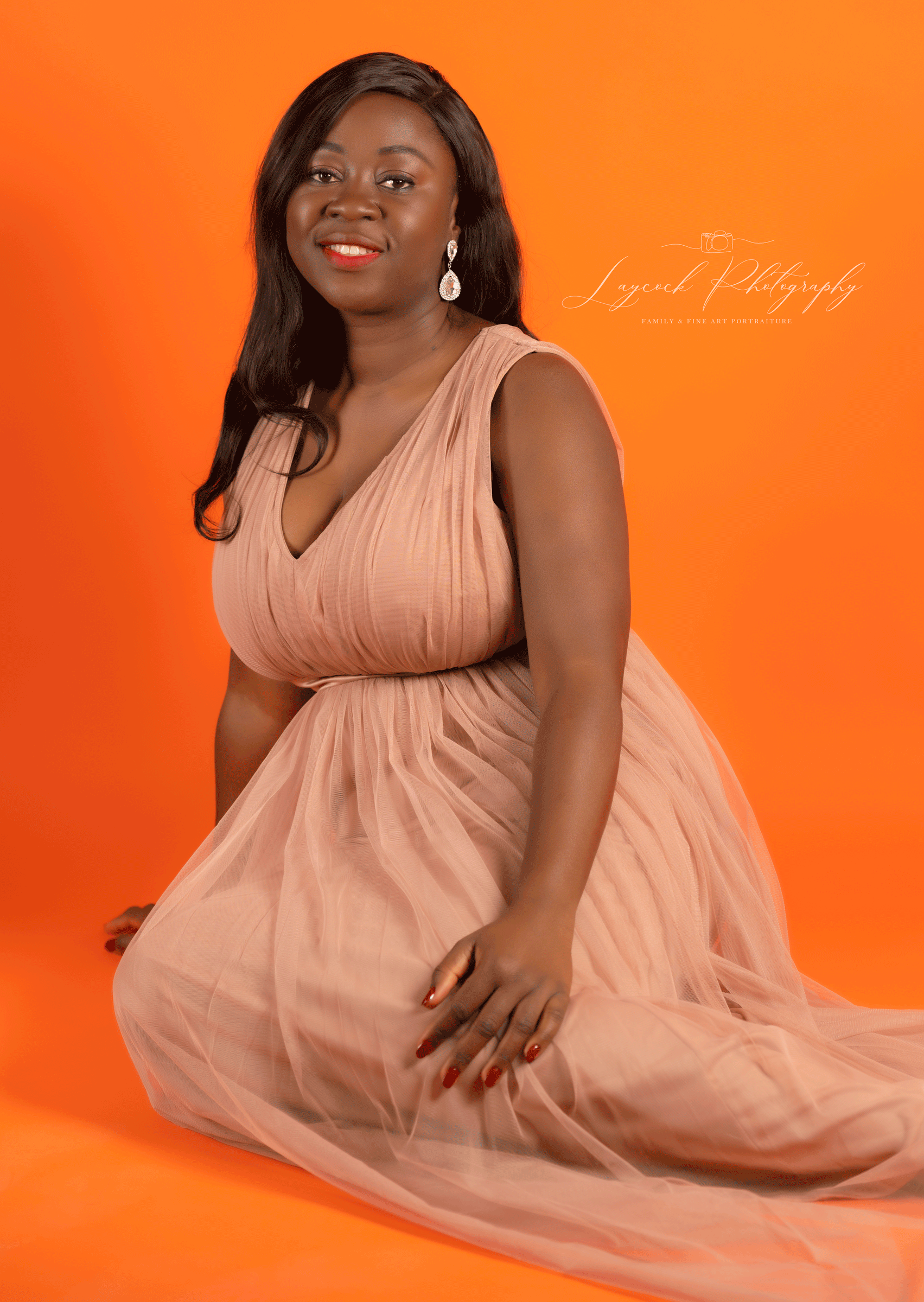 Professional studio portrait of a smiling woman wearing an elegant blush pink gown against a vibrant orange background at Laycock Photography.