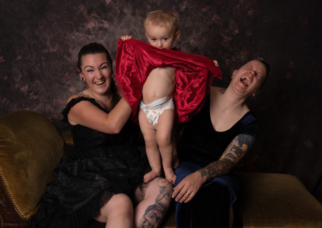 Two mothers sat on a chaise longue with their daughter wearing a red dress with it lifted up