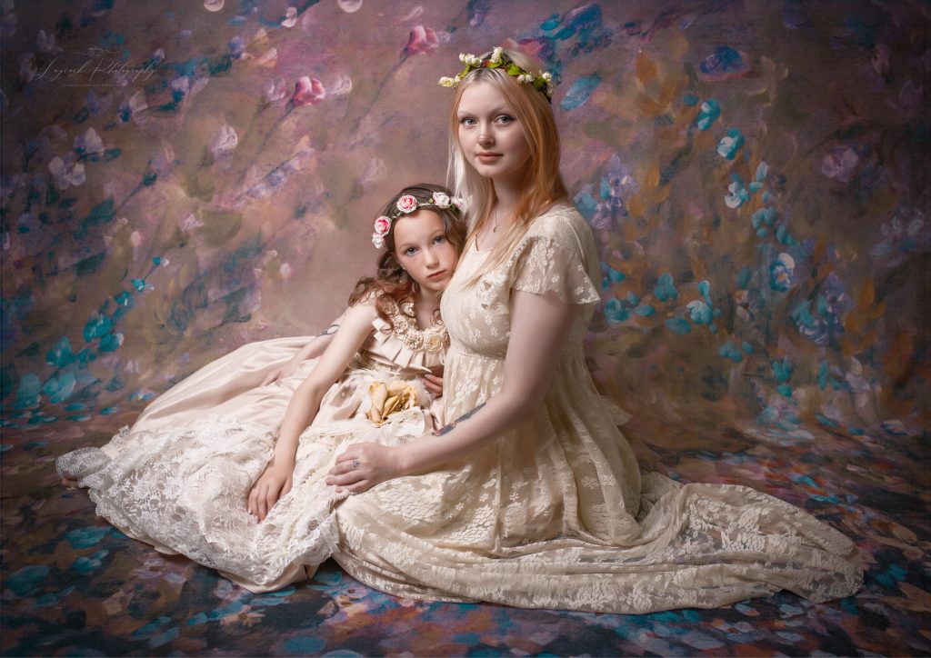 Fine art family portrait of a mother and daughter wearing elegant cream lace dresses and floral crowns, posed together against a hand-painted floral backdrop in a timeless studio portrait by Laycock Photography in Stalybridge, Greater Manchester.